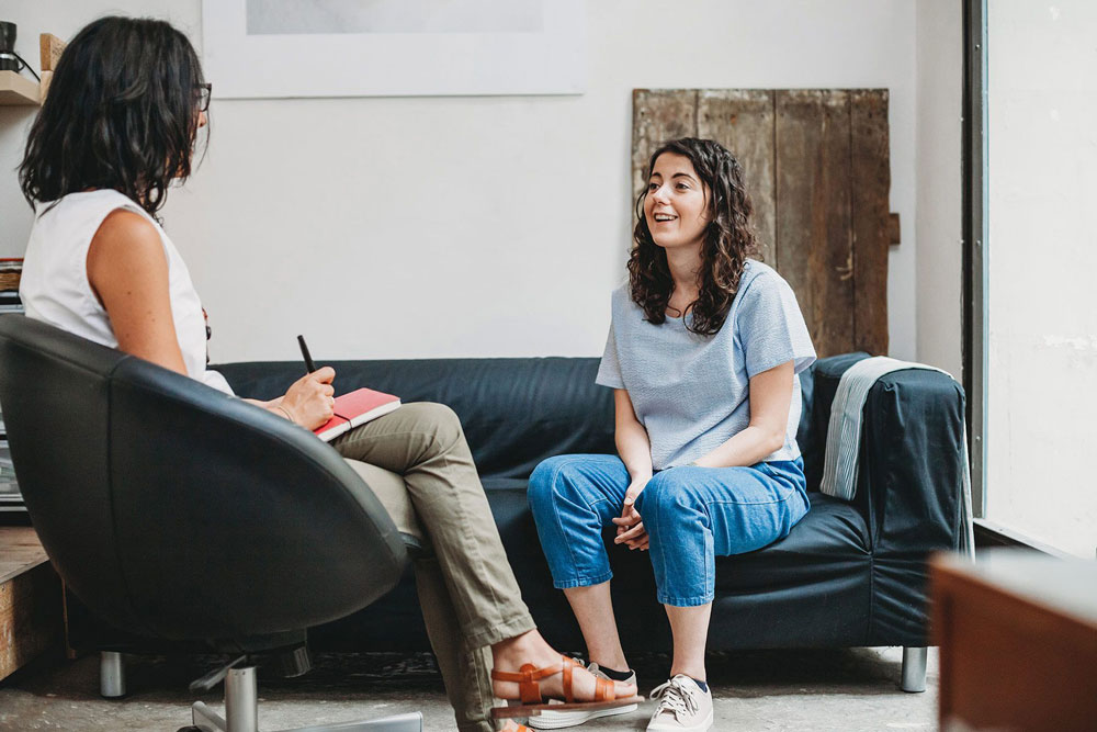 A woman sits on a couch and speaks with a female therapist.