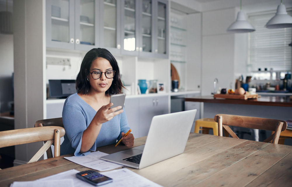 woman sitting at kitchen table with laptop and cell phone