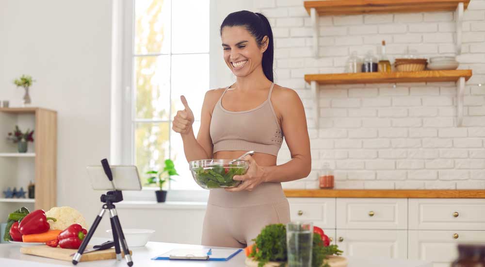 Cheerful fit woman in kitchen with smartphone on tripod holding salad bowl 